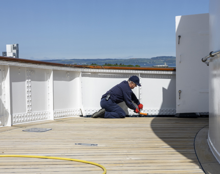 Kneeling on the deck, a Maintenance man is using a wire brush to remove paint. 