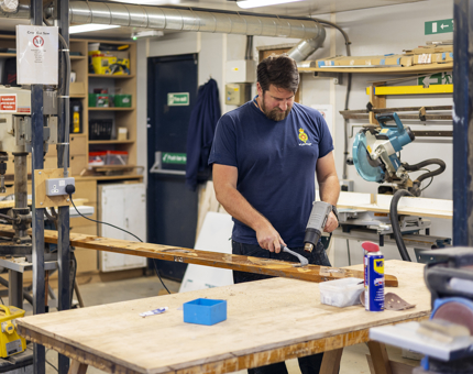 A Maintenance team member is using a heat gun to remove varnish from a plank of wood. 