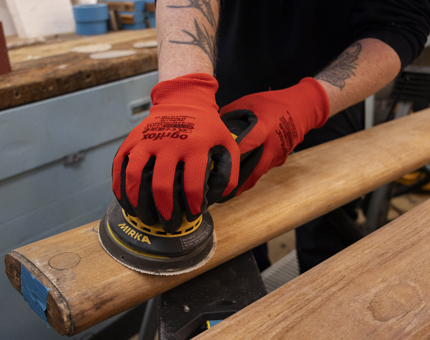 A close up of hands, wearing red gloves, holding an electric sander, sanding wooden handrails. 