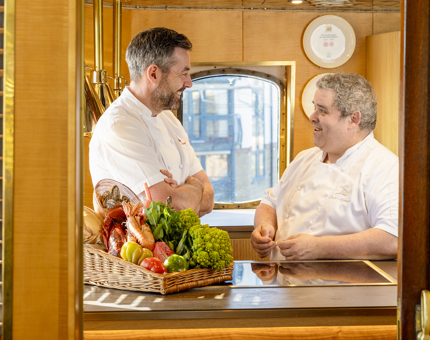 Two chefs in the Bridge's Galley aboard Fingal Hotel. There is a basket of produce on the counter. 