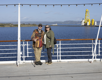 Two people posing for a photo on Britannia's Verandah Deck. There is a view of the Port of Leith in the background. 