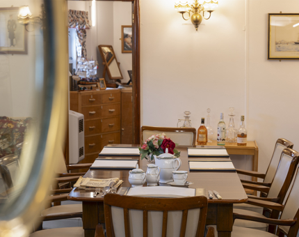 Looking through a window, a table with 8 chairs can be seen, In the background you can see through a doorway into another room. 