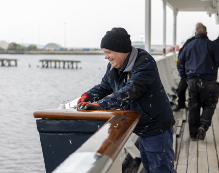 A maintenance team member holding a paint roller, applying a coat of white paint to the Starboard waist deck. You can see the Porth of Leith and other team members in the background. 