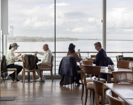 A view of people sitting at tables in the Royal Deck Tearoom. There are large picture windows showing views over the waterfront. 