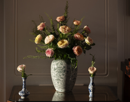 A large vase with two small bud vases is on a table. They have yellow, peach and pink flowers in them. 