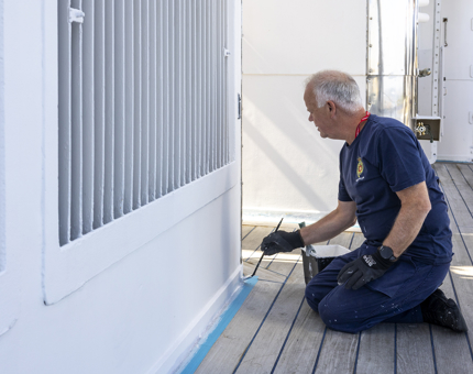 A maintenance man is kneeling on the deck painting a skirting board white. 