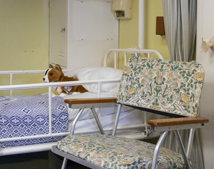 Britannia's Sick Bay. There is a hospital bed with a corgi plush toy on it and a chair next to the bed. 