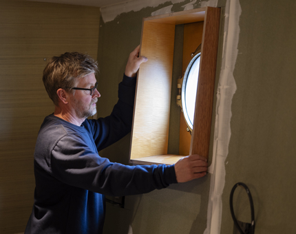 A man is fitting a wooden surround to a porthole. 