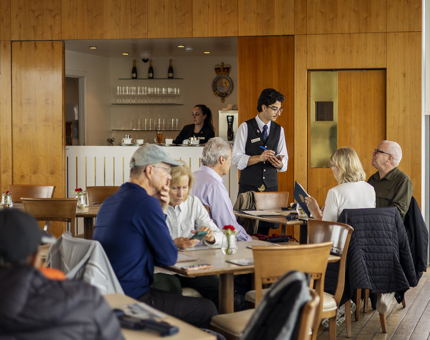 Visitors sitting at the tables in the Royal Deck Tearoom, a Hospitality Server is writing down the order for some guests. 