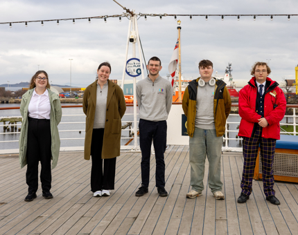 Five people standing on the Verandah Deck of The Royal Yacht Britannia in the Port of Leith. 
