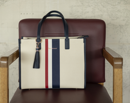 A cream handbag with a blue, red and white stripe on it. It had been placed on a chair. 