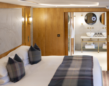 In a cabin aboard Fingal, a bed is made with white sheets and tartan blanket and pillows. You can see the bathroom through the doors in the background. 