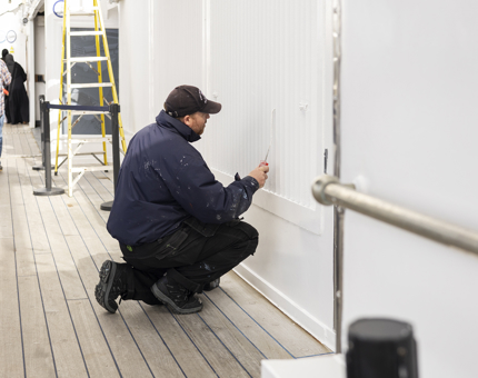 A maintenance man kneeling on the deck to apply a coat of paint. 