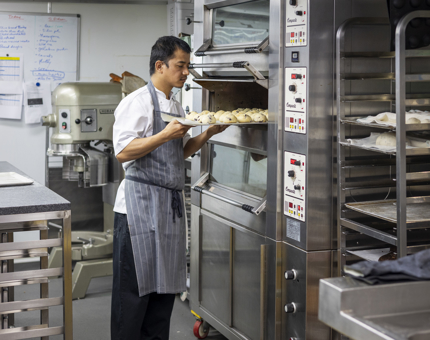 A Chef is taking a tray of bread rolls out of the oven in the Galley. 