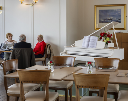 Visitors sit at a table in the Royal Deck Tearoom, there is a white baby grand piano. 