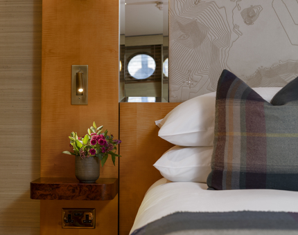 A detailed look at a bed and pillow with tartan cover on it. The headboard is great with a linear map on it. A vase of flowers is on the table next to it. 