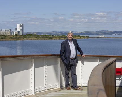 A man is posing for a photo on Britannia, the view behind him is the Firth of Forth. 