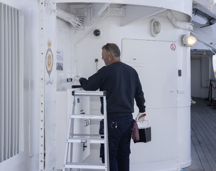 A Maintenance man is standing on the Starboard side of the Yacht, adding a coat of white paint to the wall. 