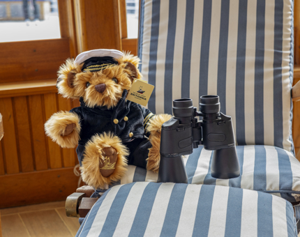 A teddy bear wearing a Captain's uniform and a pair of binoculars on a blue and white striped deck chair in the Sun Lounge. 