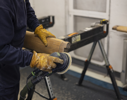 Hands wearing yellow gloves, holding an electric sander, sanding a wooden handrail. 