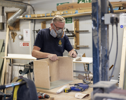 A maintenance man is in the workshop sanding a printer shelf for the Tearoom office
