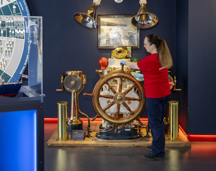A Housekeeper is in the Visitor Centre in Leith, they are dusting a ships wheel. 
