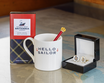 A bar of Britannia branded chocolate, a white mug with 'Hello Sailor' written on it and a crown pen inside it and cufflinks with Britannia crest on them. 