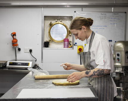 A Chef in the Galley is rolling out dough to make bread. 