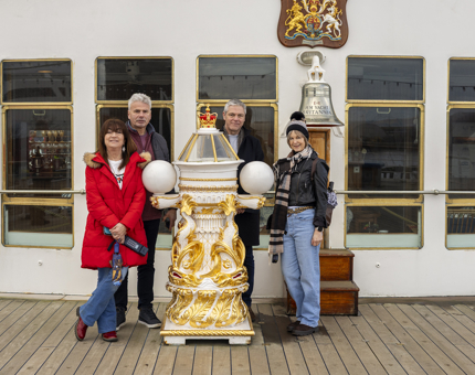 Five people posing for a photo at The Royal Yacht Britannia's decorative binnacle and ship's Bell on the Verandah Deck. 