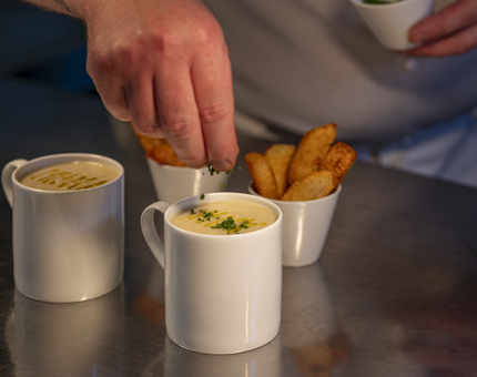 A close up of a hand sprinkling chives on a mug of soup. There is a bowl of wedges and another mug of soup on the bench too.