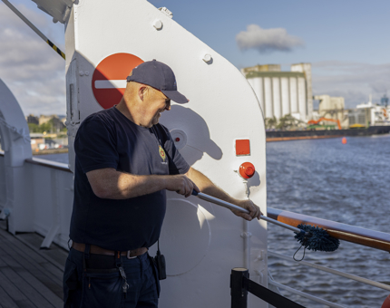 A man wearing a navy blue cap is holding a duster and is dusting the handrails on the decks of The Royal Yacht Britannia. 