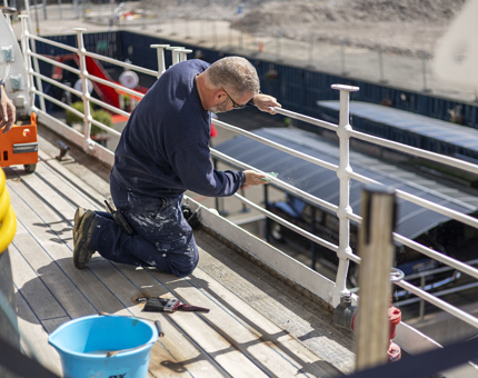 A man kneeling on the deck preparing white railings on the ship to be painted.