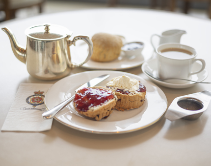 In the Tearoom there is a fruit scone with jam and cream on it, along with a silver tea pot, cup of tea and plain scone. 