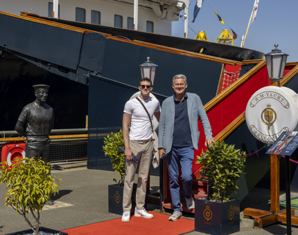 2 visitors posing for a photo at Britannia's Royal Brow. There is a statue of a sailor next to them. 