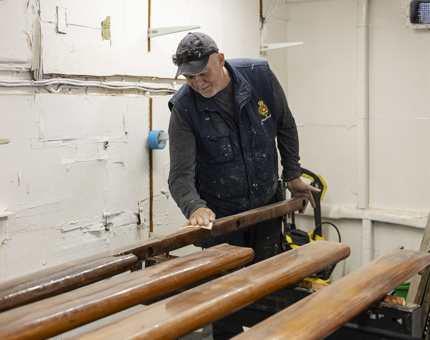 A Maintenance man is in the workshop, sanding wooden handrails. 