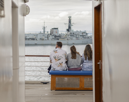 Three people sitting on a bench aboard The Royal Yacht Britannia watching a ship in the Port of Leith. 