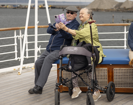 Two people sitting on a bench aboard The Royal Yacht Britannia's Verandah Deck. They are taking a selfie. 
