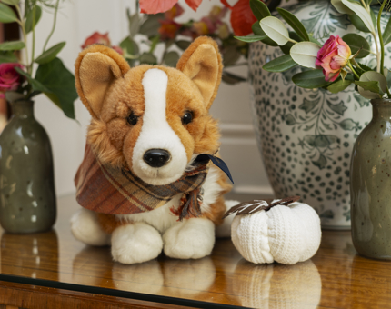 A corgi plush toy sitting on a table and is dressed in autumnal outfit. there are vases with flowers surrounding it and a small white pumpkin decoration. 