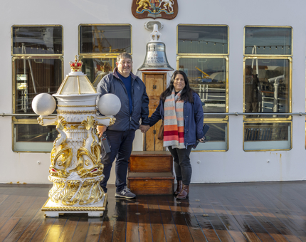 A man and woman pose for a photo by Britannia's Bell on the Verandah Deck in the Port of Leith. 