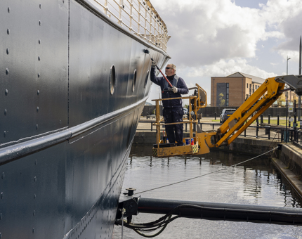 A maintenance man is on a yellow cherry picker next to Fingal Hotel, he is applying a coat of paint. 