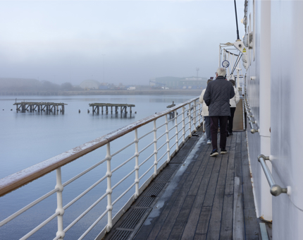 Three visitors walking along a deck aboard The Royal Yacht Britannia. The water of the Port of Leith is in the background it is foggy. 