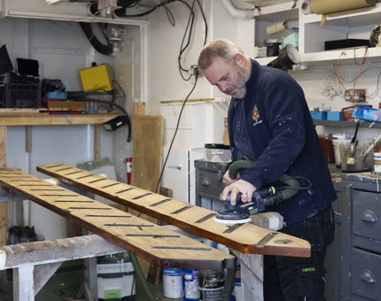 A Maintenance team member holding an electric sander to remove varnish from a wooden ladder. 