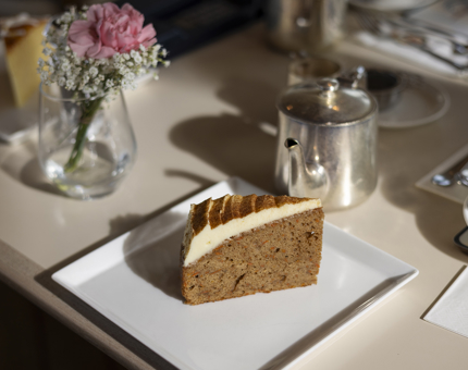 A slice of carrot cake on a table with a small silver teapot and a vase with a pink carnation. 