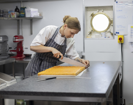 A Chef is in the Galley at a bench, cutting a slab of cake. 
