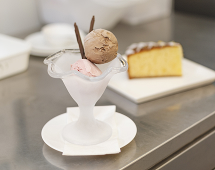 A scoop of strawberry and chocolate ice creams in a bowl. There is a slice of lemon drizzle cake in the background.