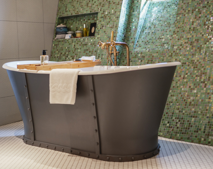 A grey roll-top bath in a cabin's bathroom aboard Fingal Hotel. The bathroom has a wall that is tiles with multicoloured small green tiles. 