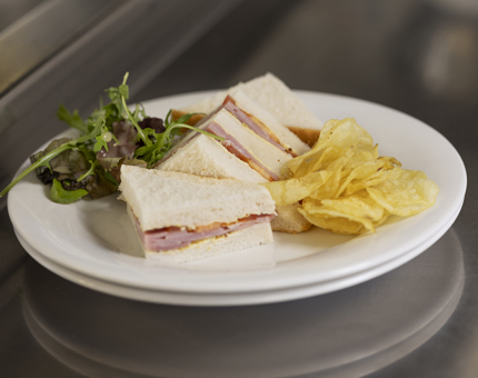A honey roast Ayrshire ham, grain mustard, vine tomato sandwich on a plate with crisps and salad, waiting to be served.