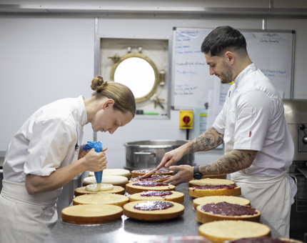 Two Chefs in the Galley putting layers of cream and jam onto layers for a Victoria sponge cake. 