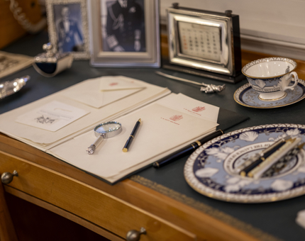 A close up of Queen Elizabeth II's desk aboard Britannia. There is headed stationery, a plate and tea cup and photographs. 