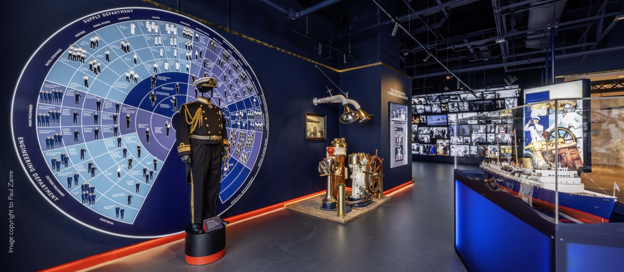 Inside the brand new Visitor Centre at Britannia. The Admiral's uniform is displayed in front of a wall display of a wheel that details all of the different job roles aboard. To the right is a model of Britannia and in the background is a cascading wall of images and video. 